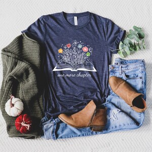 May include: A navy blue t-shirt with a white graphic of an open book with flowers growing out of it. The text "one more chapter" is printed below the book.
