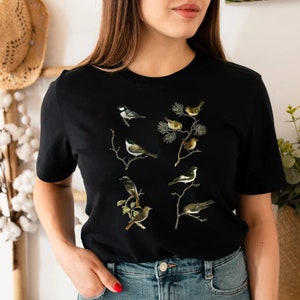 May include: Black t-shirt featuring a detailed illustration of various birds perched on branches. The birds are depicted in shades of gray, brown, and white, with intricate details. The design is centered on the front of the shirt.