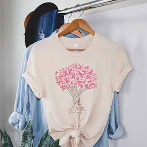May include: A cream-colored short-sleeve t-shirt featuring a graphic of a tree made of pink awareness ribbons. The tree trunk contains the words cure, hope, faith, grace, future, and strength. The shirt is tied in a knot at the bottom hem.