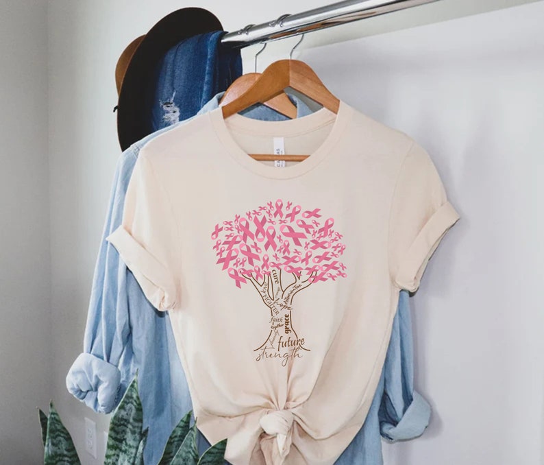 May include: A cream-colored short-sleeve t-shirt featuring a graphic of a tree made of pink awareness ribbons. The tree trunk contains the words cure, hope, faith, grace, future, and strength. The shirt is tied in a knot at the bottom hem.
