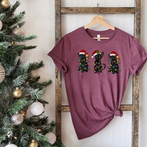 Pode incluir: Uma camiseta vinho com um desenho de tr&ecirc;s gatos pretos usando chap&eacute;us de Papai Noel e luzes de Natal.