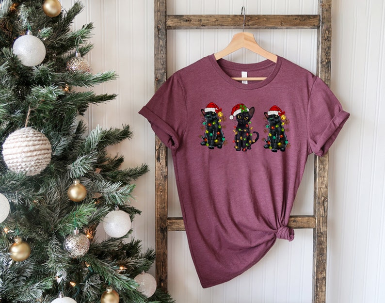 Pode incluir: Uma camiseta vinho com um desenho de tr&ecirc;s gatos pretos usando chap&eacute;us de Papai Noel e luzes de Natal.