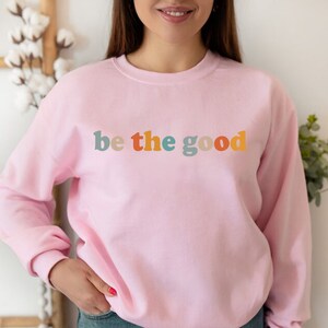 May include: A light pink sweatshirt with the phrase "be the good" in colorful, retro-style lettering across the chest. The text uses a palette of teal, orange, yellow, and blue. The sweatshirt has a crew neck and long sleeves.