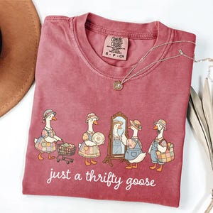 May include: A maroon Comfort Colors t-shirt featuring a whimsical illustration of geese dressed in vintage attire. The geese are depicted with shopping carts, plates, and a mirror, with the text "just a thrifty goose" below. A gold necklace and a brown hat are also visible.