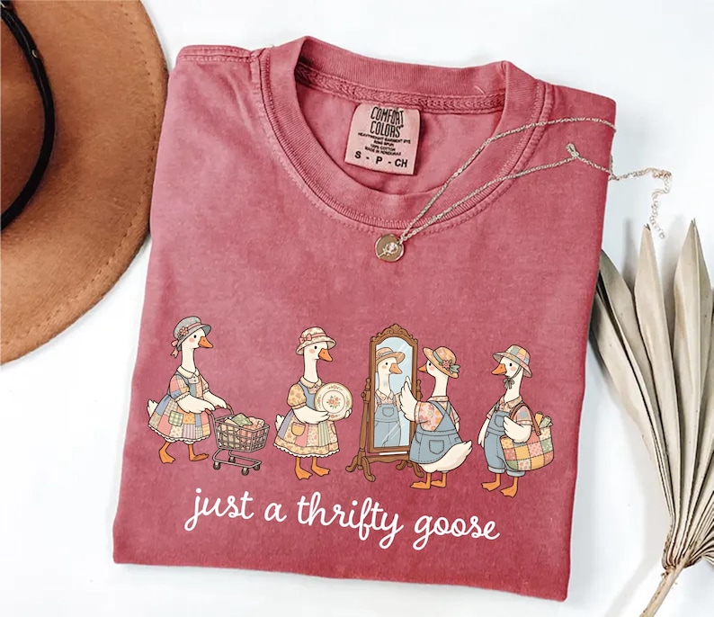 May include: A maroon Comfort Colors t-shirt featuring a whimsical illustration of geese dressed in vintage attire. The geese are depicted with shopping carts, plates, and a mirror, with the text "just a thrifty goose" below. A gold necklace and a brown hat are also visible.