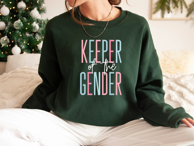May include: A dark green sweatshirt with the text "KEEPER of the GENDER" in pink, blue, and white lettering.