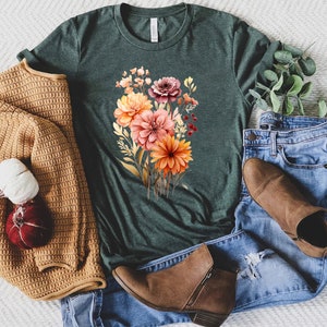May include: A dark forest green short-sleeve t-shirt featuring a central graphic of a colorful floral bouquet with orange, pink, and deep red blossoms, paired with a mustard yellow knit cardigan, blue denim jeans, and brown ankle boots.