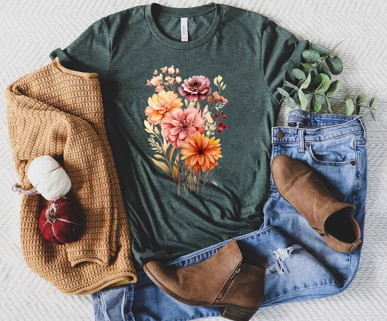 May include: A dark forest green short-sleeve t-shirt featuring a central graphic of a colorful floral bouquet with orange, pink, and deep red blossoms, paired with a mustard yellow knit cardigan, blue denim jeans, and brown ankle boots.
