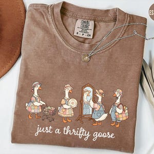 May include: Espresso-colored t-shirt with a whimsical illustration of geese dressed in vintage attire. The geese are depicted shopping, admiring themselves in a mirror, and carrying baskets. The text "just a thrifty goose" is printed below the illustration. A silver necklace is draped across the shirt.