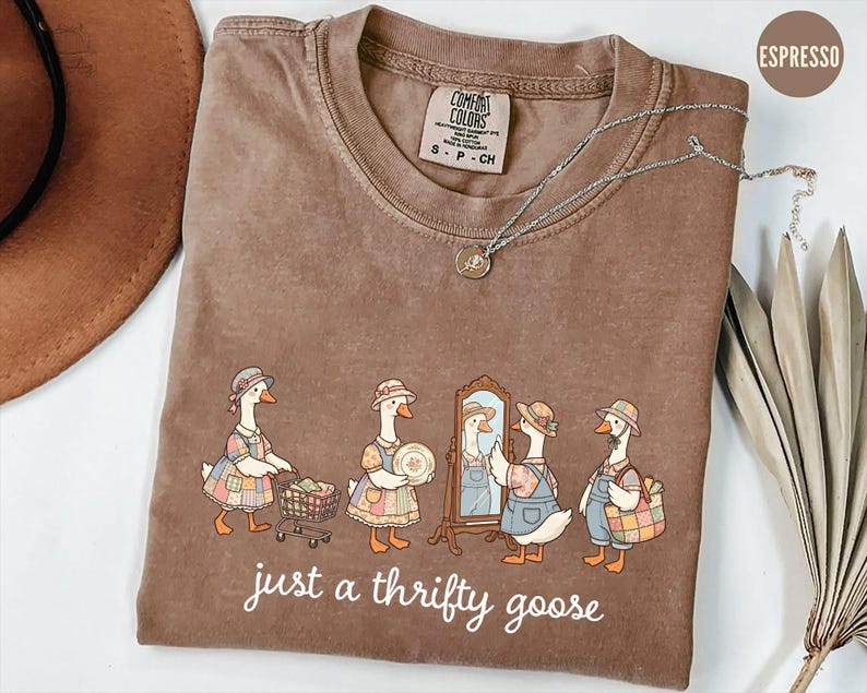 May include: Espresso-colored t-shirt with a whimsical illustration of geese dressed in vintage attire. The geese are depicted shopping, admiring themselves in a mirror, and carrying baskets. The text "just a thrifty goose" is printed below the illustration. A silver necklace is draped across the shirt.