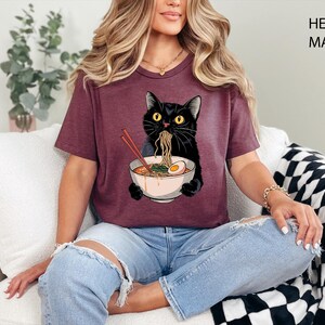 May include: A heather maroon t-shirt featuring a black cat eating ramen noodles from a bowl. The cat is wearing a white collar and has a surprised expression. The text "HEATHER MAROON" is visible in the upper right corner of the image.