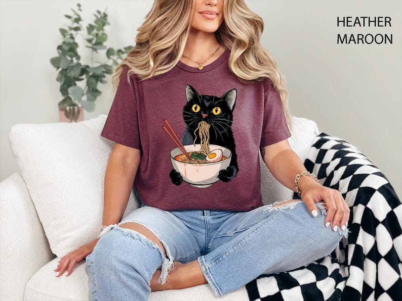 May include: A heather maroon t-shirt featuring a black cat eating ramen noodles from a bowl. The cat is wearing a white collar and has a surprised expression. The text "HEATHER MAROON" is visible in the upper right corner of the image.