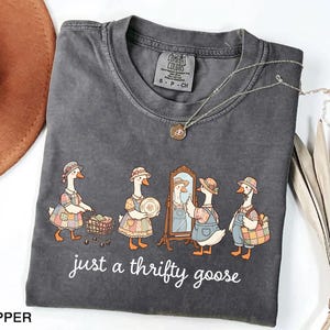 May include: Dark gray Comfort Colors t-shirt featuring a whimsical illustration of geese dressed in vintage clothing. The geese are depicted shopping, admiring themselves in a mirror, and carrying a basket. The text "just a thrifty goose" is printed below the illustration.