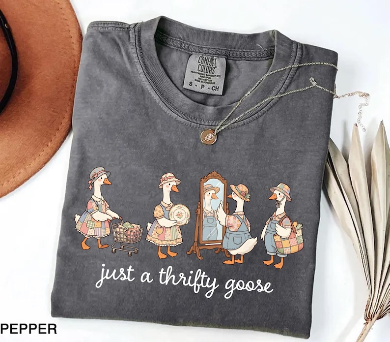 May include: Dark gray Comfort Colors t-shirt featuring a whimsical illustration of geese dressed in vintage clothing. The geese are depicted shopping, admiring themselves in a mirror, and carrying a basket. The text "just a thrifty goose" is printed below the illustration.