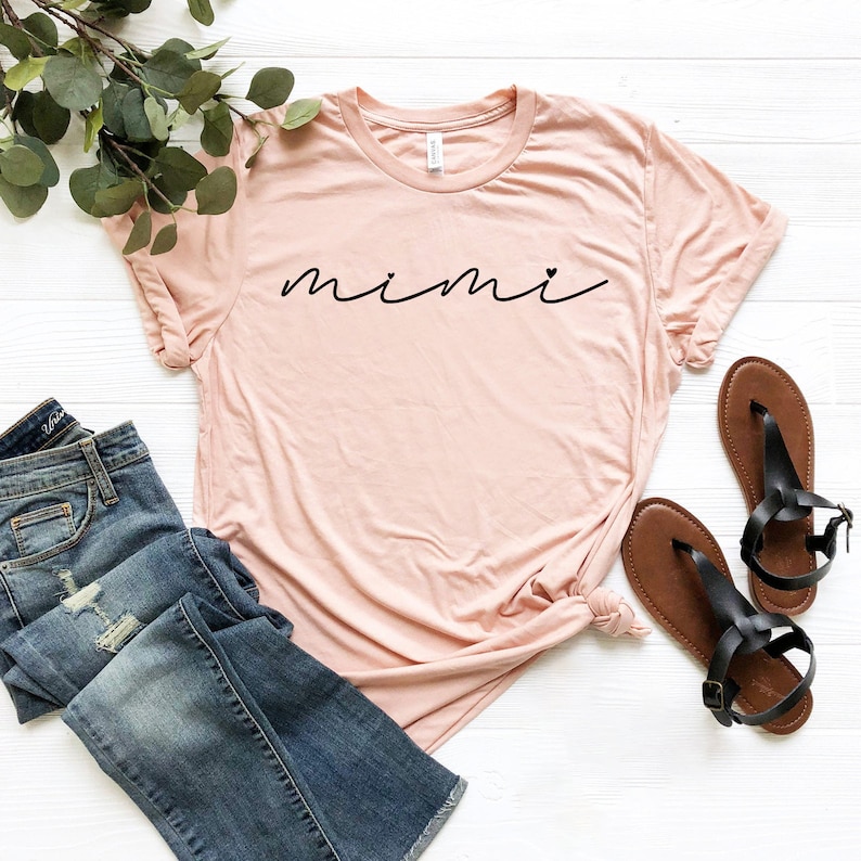 May include: A light pink t-shirt with the word "mimi" written in black cursive script with a small heart after the "i".