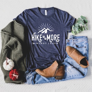 May include: A navy blue t-shirt with the text "Hike More Worry Less" in white, featuring a mountain and paw print design. The shirt is paired with blue jeans, brown boots, and a green sweater. Decorative pumpkins are also present.
