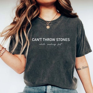 Puede incluir: Una camiseta gris oscuro con el texto "CAN'T THROW STONES while washing feet" impreso en blanco.