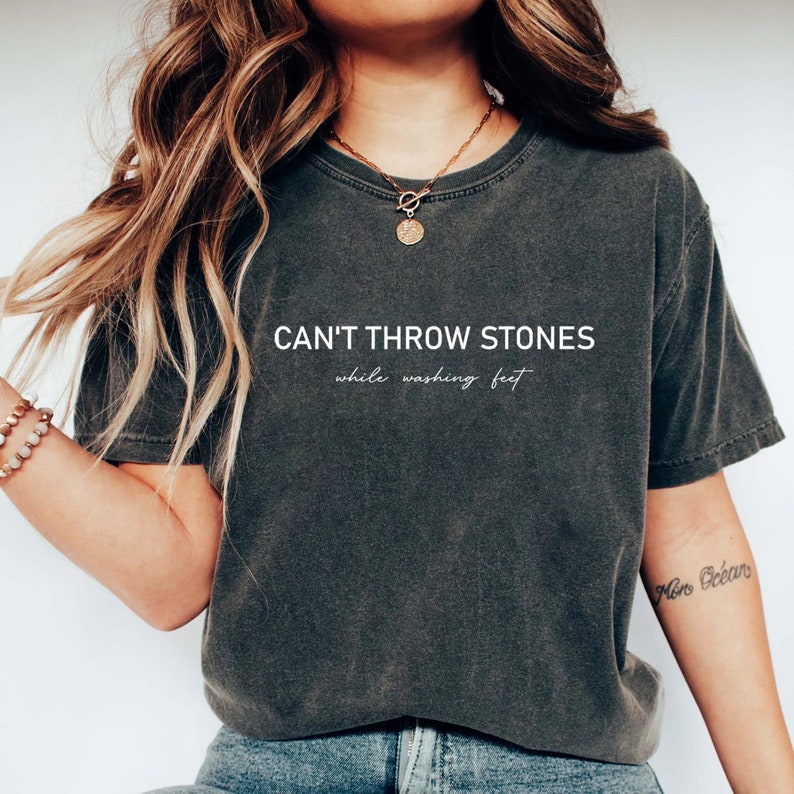 Puede incluir: Una camiseta gris oscuro con el texto "CAN'T THROW STONES while washing feet" impreso en blanco.