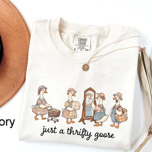 May include: Ivory-colored Comfort Colors t-shirt featuring a whimsical illustration of geese shopping. The geese are dressed in vintage clothing, with the text "just a thrifty goose" in a handwritten font. A gold necklace and a brown hat are also visible.