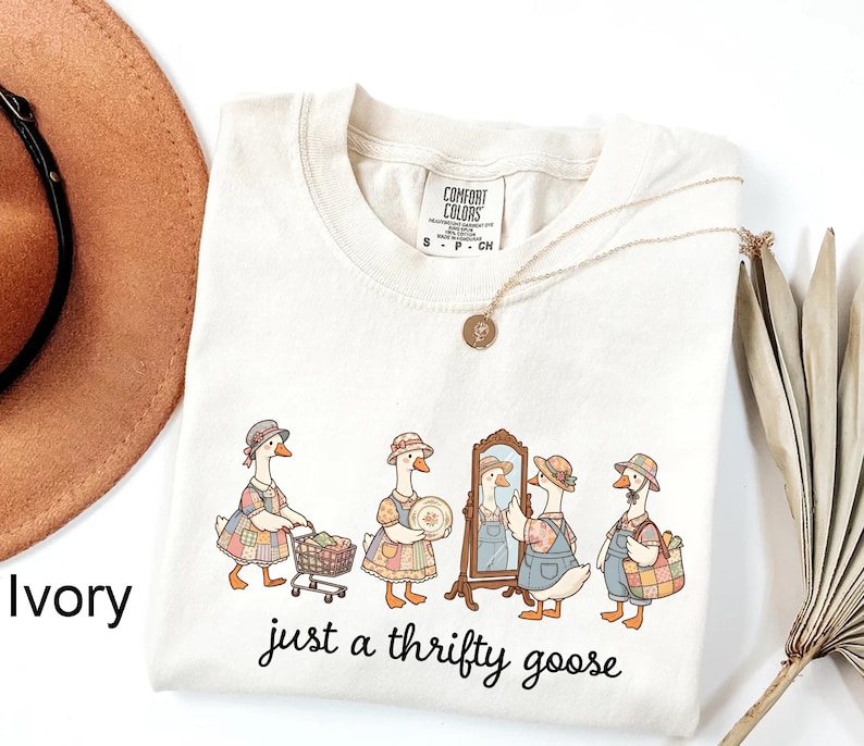 May include: Ivory-colored Comfort Colors t-shirt featuring a whimsical illustration of geese shopping. The geese are dressed in vintage clothing, with the text "just a thrifty goose" in a handwritten font. A gold necklace and a brown hat are also visible.