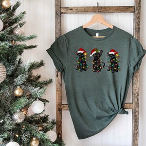 Pode incluir: Uma camiseta verde com tr&ecirc;s gatos pretos usando chap&eacute;us de Papai Noel e luzes de Natal. Os gatos est&atilde;o sentados em fila.