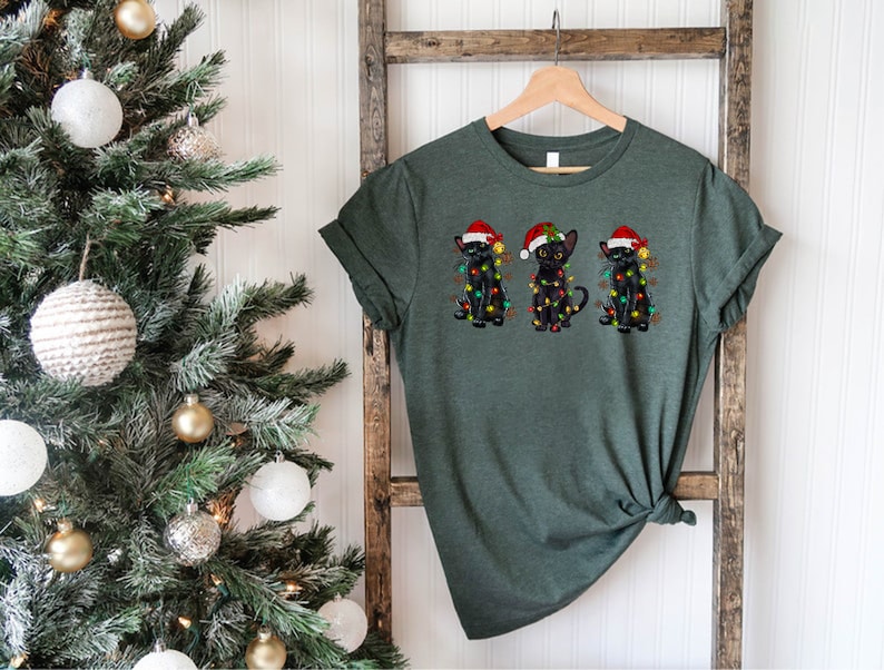 Pode incluir: Uma camiseta verde com tr&ecirc;s gatos pretos usando chap&eacute;us de Papai Noel e luzes de Natal. Os gatos est&atilde;o sentados em fila.
