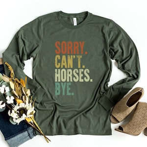May include: A green long-sleeved t-shirt with the text "SORRY. CAN'T. HORSES. BYE." printed in a vintage style.