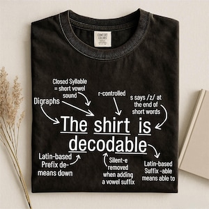 May include: A black crew neck t-shirt featuring white text that reads The shirt is decodable, with arrows pointing to linguistic labels including Digraphs, Closed Syllable, r-controlled, s says /z/, Latin-based Prefix de-, Silent-e, and Latin-based Suffix -able.