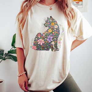 May include: Cream-colored t-shirt featuring a floral cat design. The cat is composed of various colorful flowers, including pink, purple, and yellow blooms. The shirt has short sleeves and a relaxed fit, suitable for casual wear.
