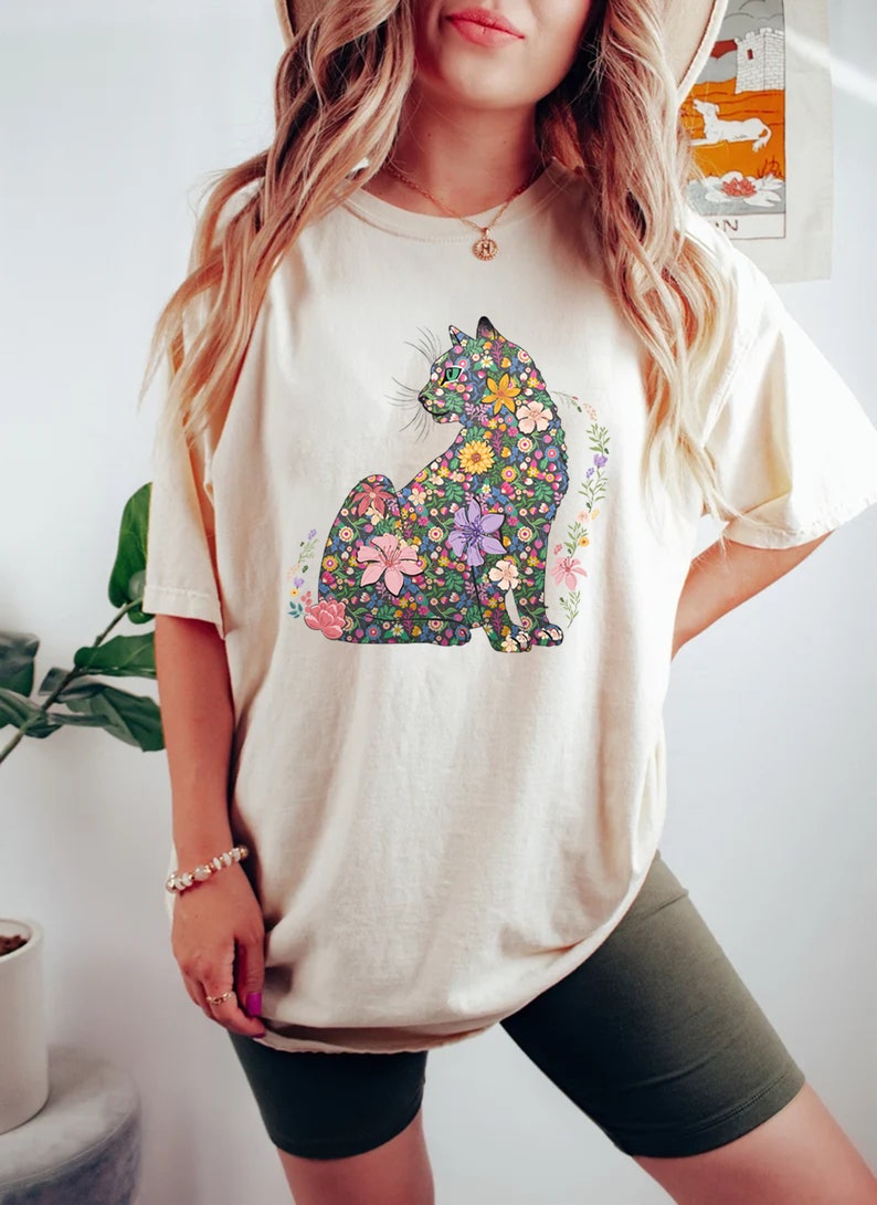 May include: Cream-colored t-shirt featuring a floral cat design. The cat is composed of various colorful flowers, including pink, purple, and yellow blooms. The shirt has short sleeves and a relaxed fit, suitable for casual wear.