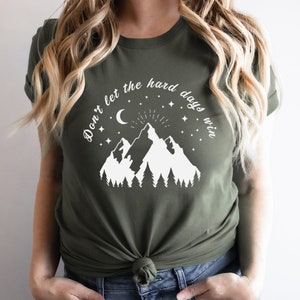 以下が含まれることがあります： オリーブグリーンのTシャツ。白いグラフィックは山、木、三日月、星。テキスト「Don't let the hard days win」は山のデザインの上にアーチ状に配置されています。柔らかい素材で作られたシャツです。