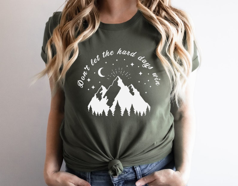 以下が含まれることがあります： オリーブグリーンのTシャツ。白いグラフィックは山、木、三日月、星。テキスト「Don't let the hard days win」は山のデザインの上にアーチ状に配置されています。柔らかい素材で作られたシャツです。