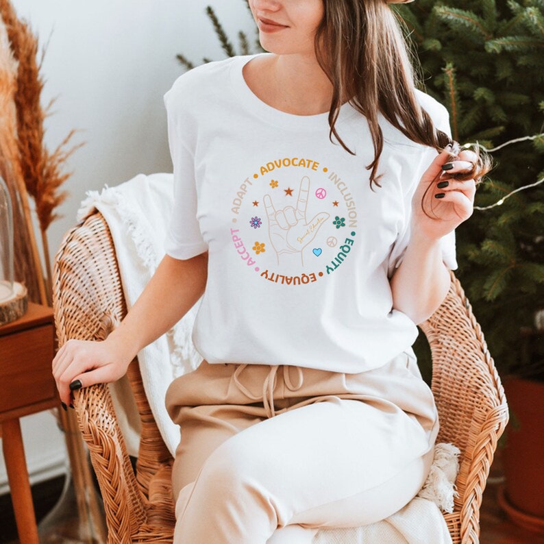 May include: White t-shirt with a colorful graphic design featuring a hand making the American Sign Language sign for "I love you". The graphic is surrounded by a circle of words: "Adapt", "Accept", "Equality", "Equity", "Advocate", "Inclusion".