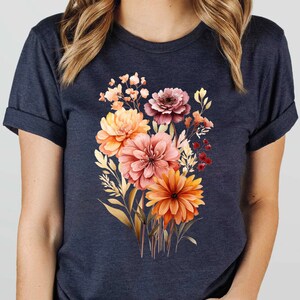 May include: A dark navy blue short-sleeve t-shirt featuring a central graphic print of a vibrant floral bouquet. The bouquet consists of various flowers in shades of orange, peach, pink, and deep red, accompanied by golden-toned foliage and stems.