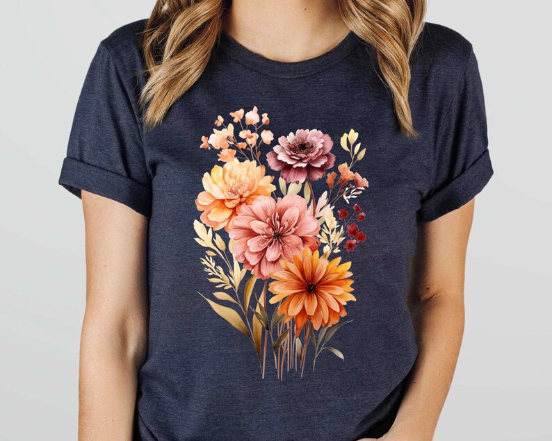 May include: A dark navy blue short-sleeve t-shirt featuring a central graphic print of a vibrant floral bouquet. The bouquet consists of various flowers in shades of orange, peach, pink, and deep red, accompanied by golden-toned foliage and stems.
