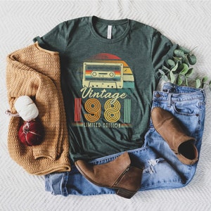May include: Green t-shirt with a retro cassette tape design and the text "Vintage 1981 Limited Edition." The shirt is paired with blue jeans, brown boots, and a tan sweater. A decorative item is also included.