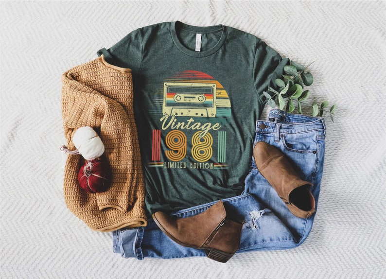 May include: Green t-shirt with a retro cassette tape design and the text "Vintage 1981 Limited Edition." The shirt is paired with blue jeans, brown boots, and a tan sweater. A decorative item is also included.