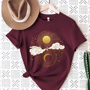 May include: A maroon t-shirt with a gold sun and moon design. The sun and moon are surrounded by clouds and stars.