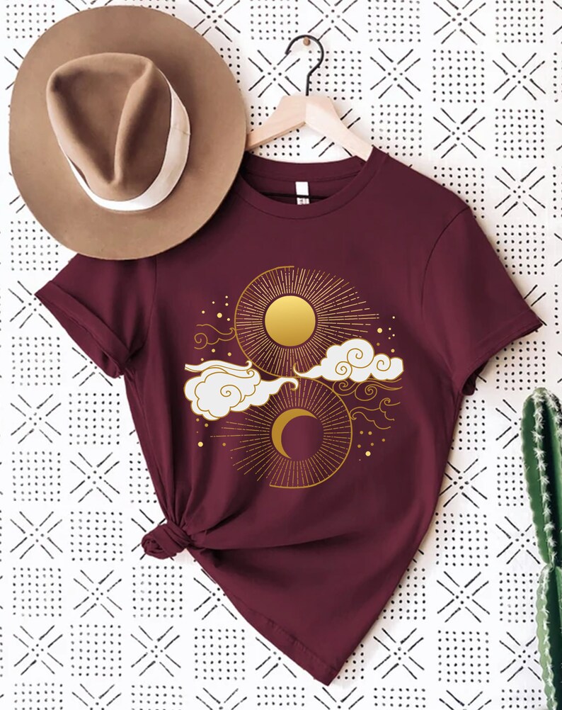 May include: A maroon t-shirt with a gold sun and moon design. The sun and moon are surrounded by clouds and stars.