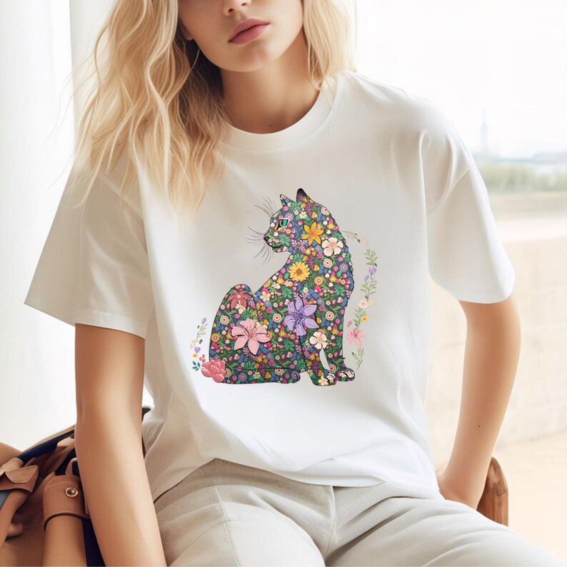 May include: White t-shirt featuring a seated cat design. The cat is intricately detailed with a floral pattern in shades of pink, purple, yellow, and green. The design is surrounded by a floral border, creating a whimsical and artistic look.
