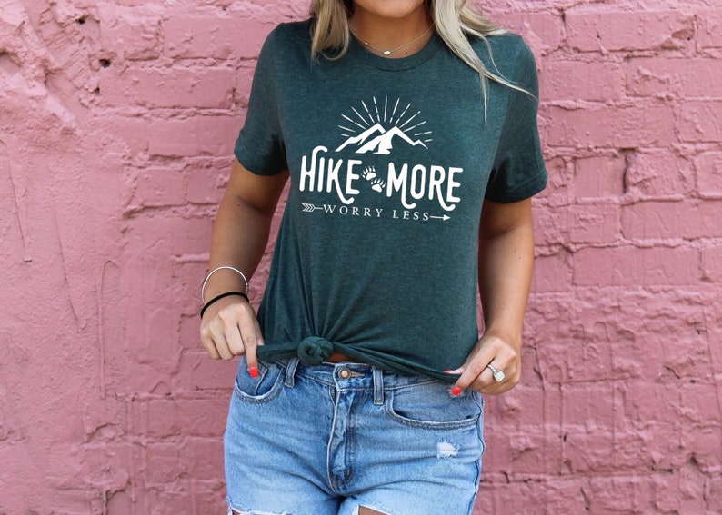 May include: Dark green t-shirt with the text "Hike More Worry Less" in white. The design includes a mountain range, sun rays, and paw prints. The shirt is knotted at the waist and paired with blue denim shorts.