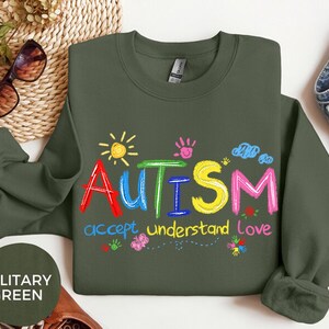 May include: Olive green sweatshirt with the word "AUTISM" in colorful letters, accompanied by the phrases "accept, understand, love." The design includes a sun, handprints, clouds, and flowers. A casual, comfortable top.