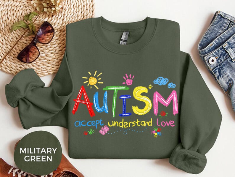 May include: Olive green sweatshirt with the word "AUTISM" in colorful letters, accompanied by the phrases "accept, understand, love." The design includes a sun, handprints, clouds, and flowers. A casual, comfortable top.