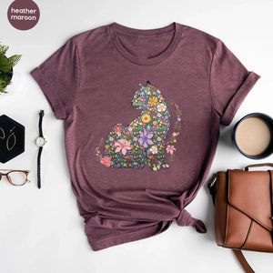 May include: Heather maroon t-shirt featuring a floral cat design. The cat is composed of various colorful flowers, including pink, yellow, and purple. The shirt has rolled-up sleeves and the text "heather maroon" is in the upper left corner.