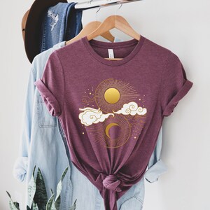 May include: A maroon t-shirt with a gold sun and moon design. The sun and moon are surrounded by clouds and stars.