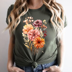 May include: An olive green short-sleeved t-shirt featuring a central graphic print of a colorful floral bouquet. The bouquet includes various flowers in shades of orange, pink, and deep red, accompanied by green and gold foliage elements.