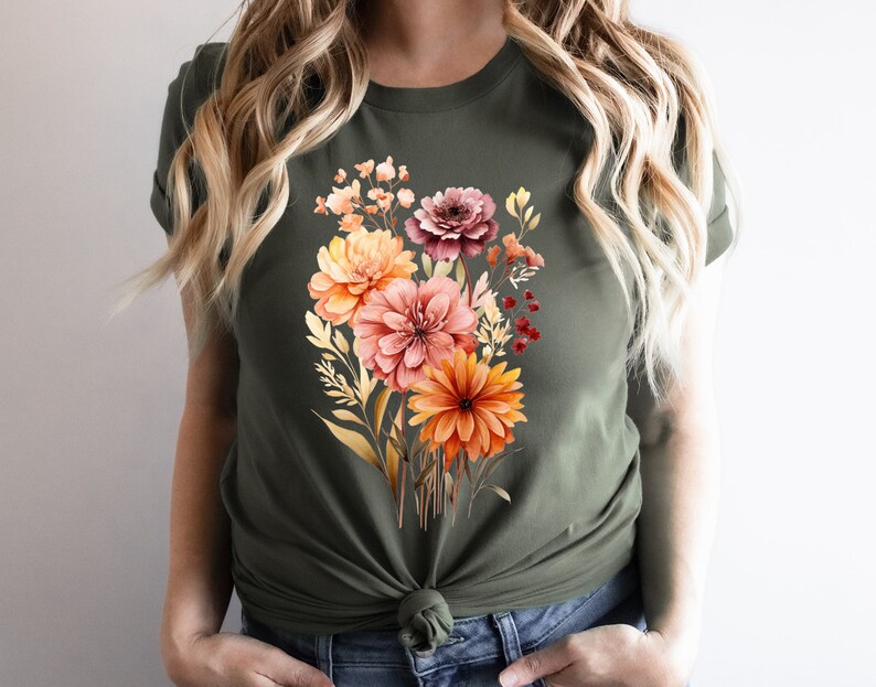 May include: An olive green short-sleeved t-shirt featuring a central graphic print of a colorful floral bouquet. The bouquet includes various flowers in shades of orange, pink, and deep red, accompanied by green and gold foliage elements.