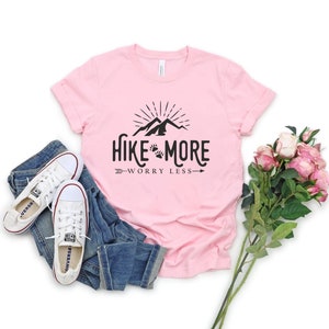 May include: A light pink t-shirt with the words "Hike More Worry Less" in black, along with a mountain and paw print design. The shirt is paired with blue jeans and white sneakers. A bouquet of pink roses is also present.