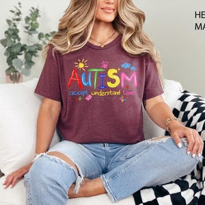 May include: Heather maroon t-shirt with the word "AUTISM" in colorful letters, with the words "accept understand love" underneath. The shirt has a crew neck and short sleeves. The model is wearing blue jeans.