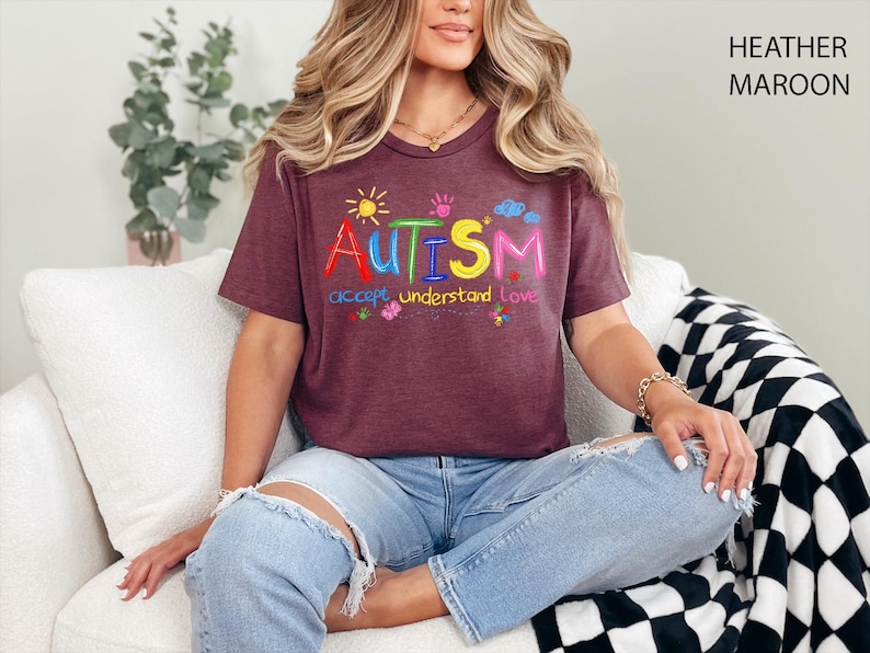 May include: Heather maroon t-shirt with the word "AUTISM" in colorful letters, with the words "accept understand love" underneath. The shirt has a crew neck and short sleeves. The model is wearing blue jeans.